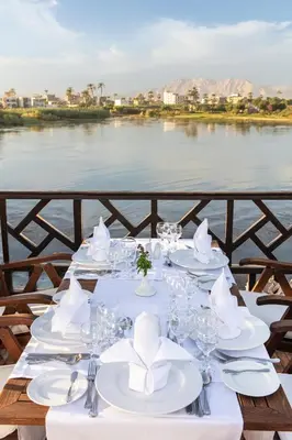 a table with white plates and napkins and a view of a river at JAZ Farouz El Nil III Nile Cruise - Every Saturday from Luxor for 07 & 05 Nights in Luxor