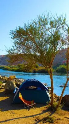 a tent and a chair under a tree next to a river at ASSINGAR camp in Jazīrat Suhayl