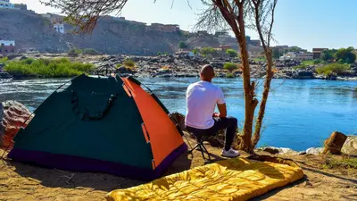 a man sitting next to a tent next to a river at ASSINGAR camp in Jazīrat Suhayl