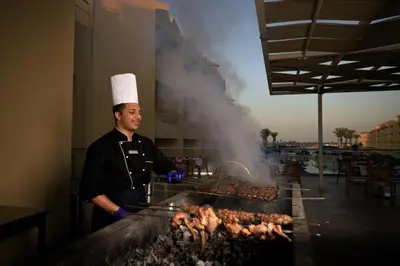 a man cooking meat on a grill with smoke at Amwaj Beach Club Abu Soma in Hurghada